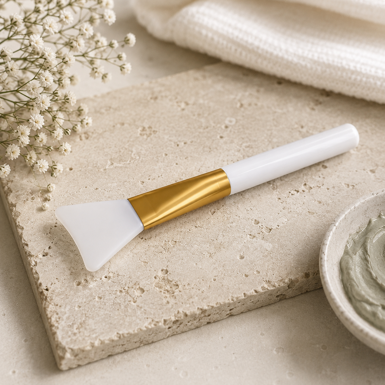 Silicone mask applicator brush with white handle and gold accent, styled on a marble tray beside a bowl of clay mask